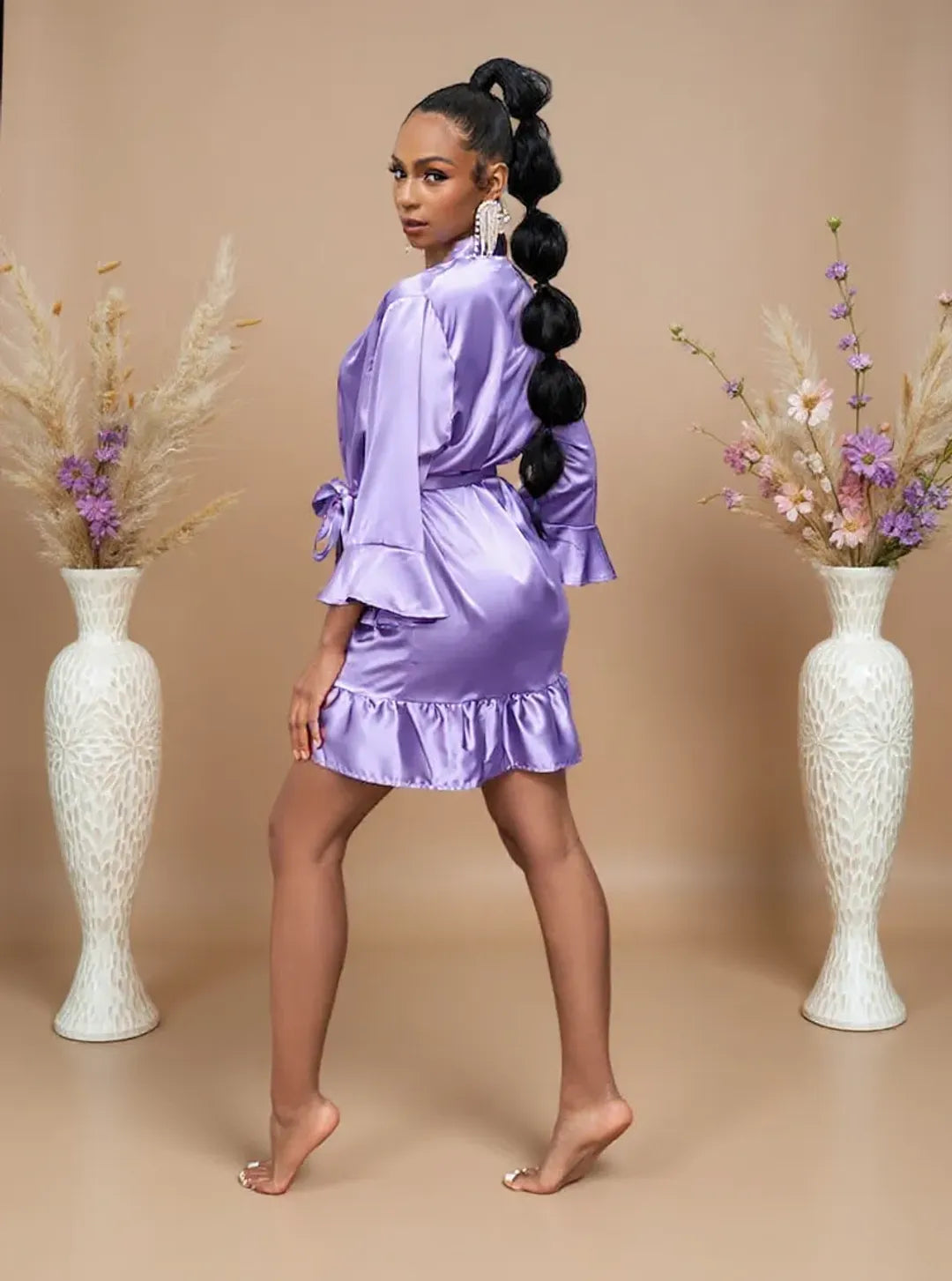 Woman in a purple satin outfit standing between two decorative vases with flowers on a beige background